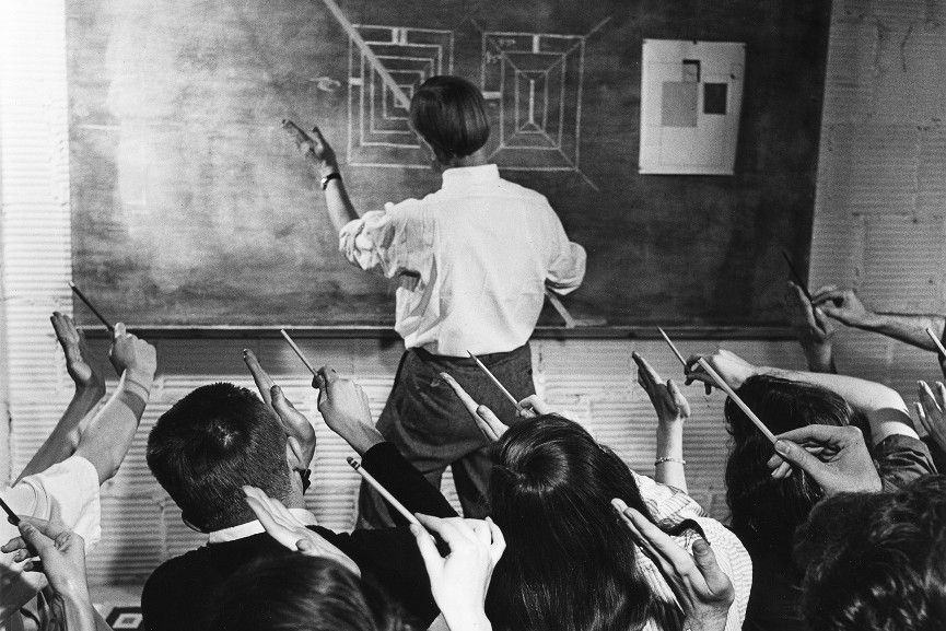 Black and white photograph showing the white shirted back of a t-shirt standing at a chalkboard with hands stretched out to the left. In foreground multiple heads and hands of students holding pencils and mimicing the angle being shown by the teacher.